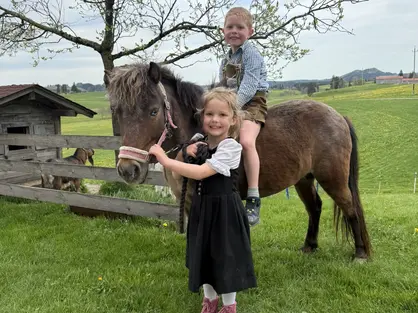 Ponyreiten auf Lotti © Nicole Stadler Zwei Kinder befinden sich mit einem braunen Pony auf einer grünen Wiese. Ein Junge sitzt in Lederhose auf dem Pony, das Mädchen steht davor und hält das Halfter. Im Hintergrund sind ein Holzzaun, ein blühender Baum, ein Ziegenhäuschen und sanfte Hügel zu sehen.