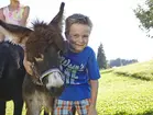 Eselumarmung © www.westenried.de Ein Junge steht auf einer grünen Wiese und umarmt einen Esel; im Hintergrund ist ein weiterer Esel und Bäume zu sehen.