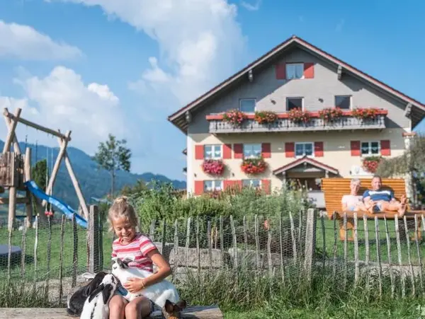 Mädchen sitzt im Garten mit einer Ziege. Im Hintergrund ein Bauernhof mit Spielplatz und den Eltern im Liegestuhl