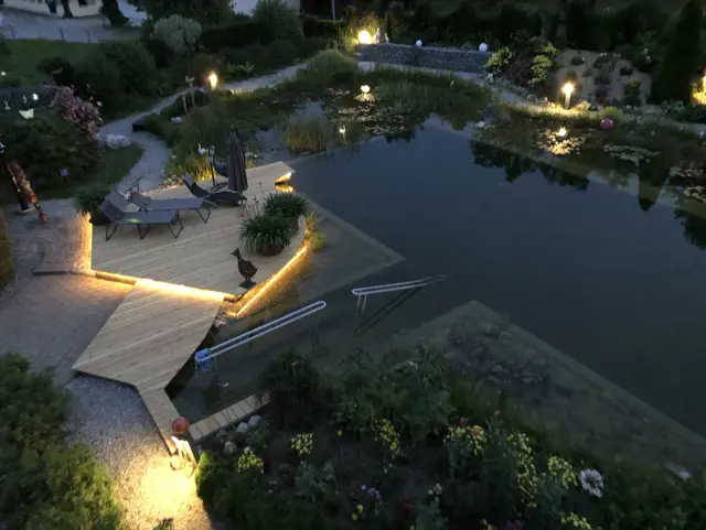 Abendliche Außenanlage im Allgäu mit beleuchtetem Naturschwimmteich, Wegen und Sitzbereichen in idyllischer Urlaubsatmosphäre.