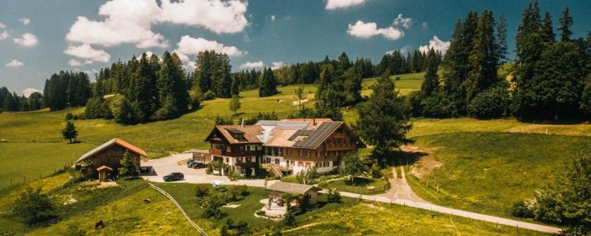 Luftaufnahme eines MirAllgäuer Bauernhofs im Allgäu, umgeben von grünen Wiesen, Bäumen und naturnaher Landschaft. Der Hof liegt idyllisch inmitten der Allgäuer Natur unter blauem Himmel.